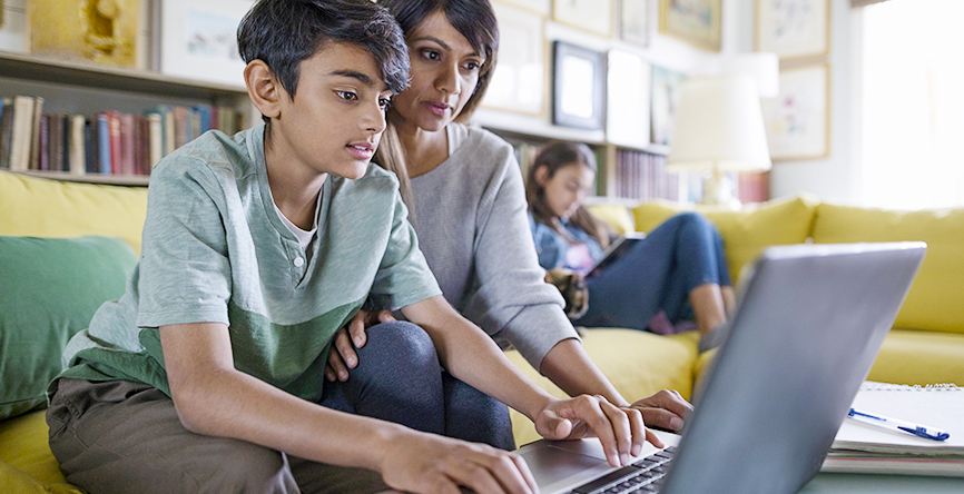 Young boy sits on a sofa with his mum whilst working on his laptop