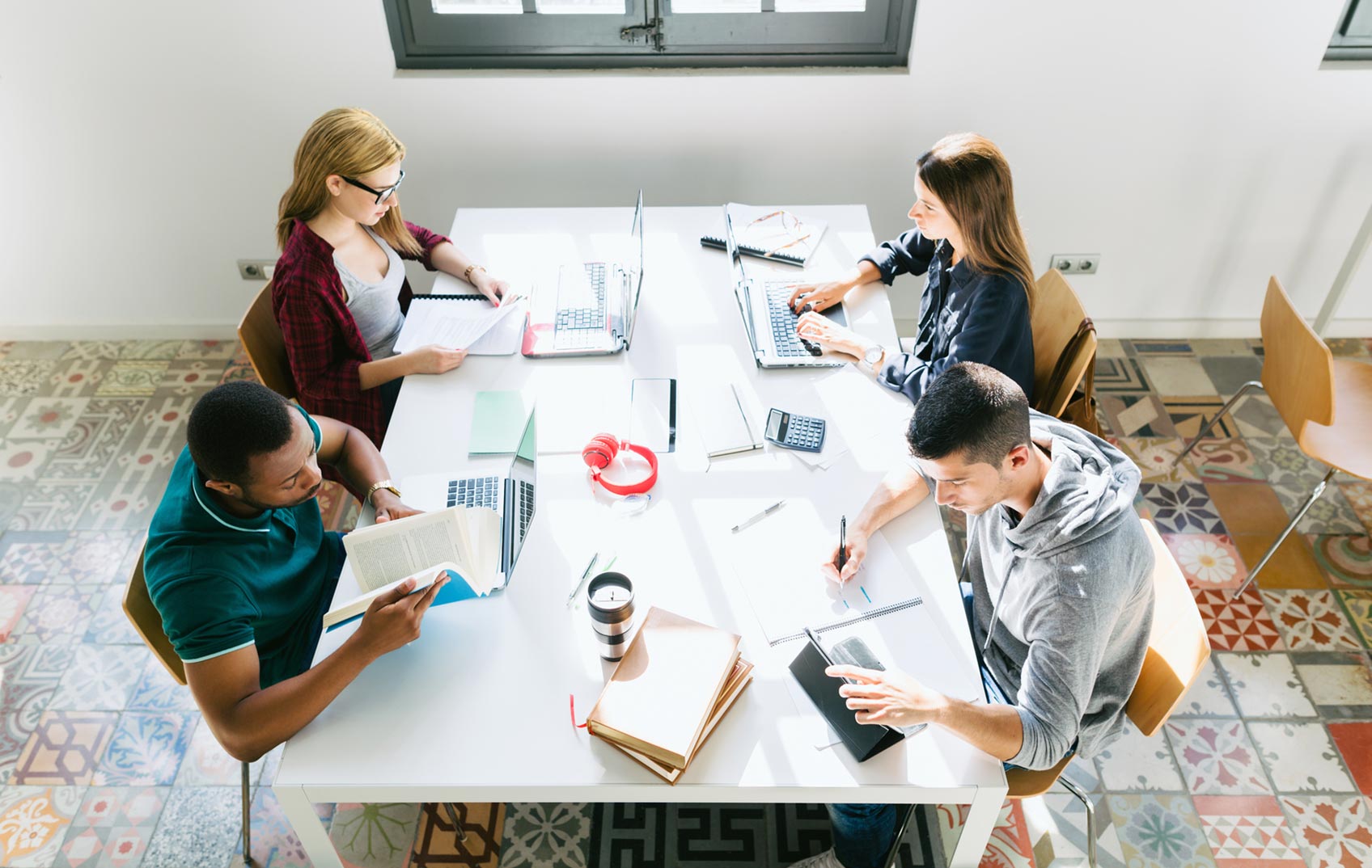 A group of young adults sit together at a table with their laptops and books studying