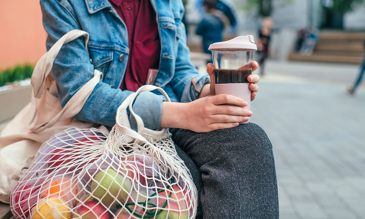 A lady sits on a bench outside wih her reusable coffee cup and resuable shopping bag full of fruit and vegetables