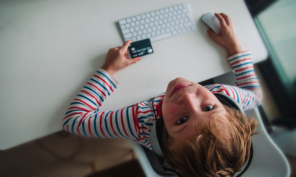 A young boy sits at his computer holding a bank card and looking upwards at the camera