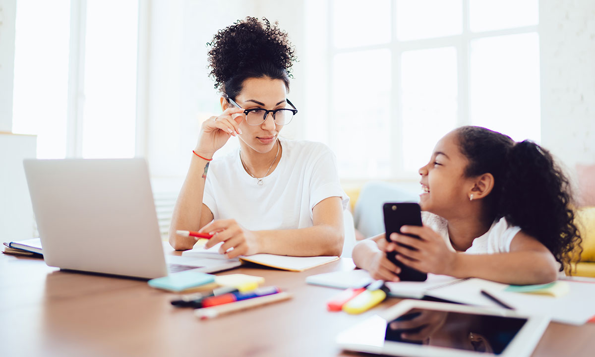 A mother and daugher sit at a table working. The young daughter giggles whilst looking at the mobile she is holding