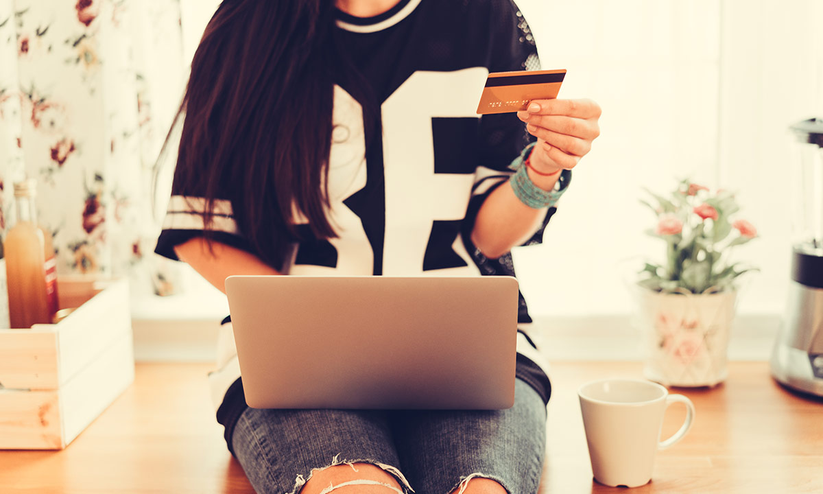 A teenage girl sits on the kitchen worktop with her laptop and bank card in her hands. 