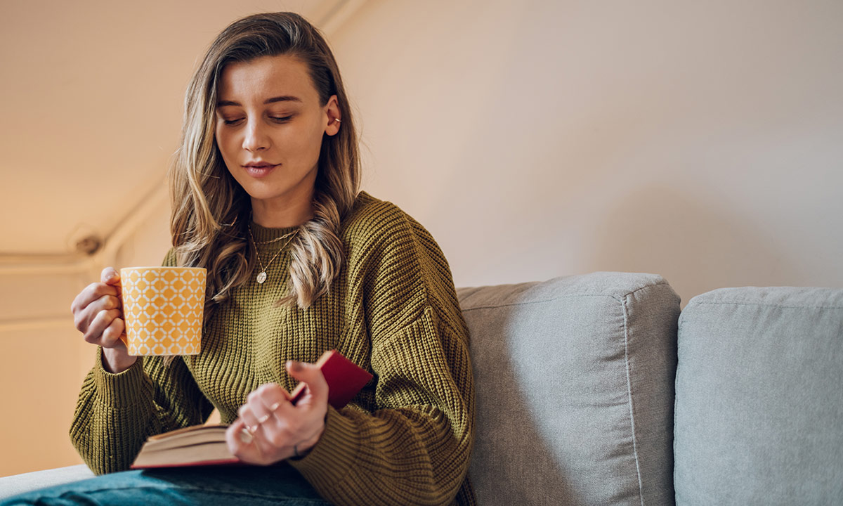 A teenage girl sits on the sofa with a mug of tea whilst reading a book