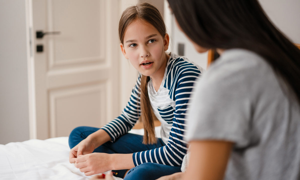 A young girl sits cross legged on her bed whilst chatting with her mum